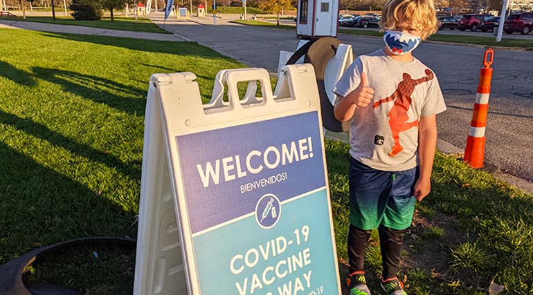 Child in face mask holding thumbs up next to COVID-19 vaccine sign